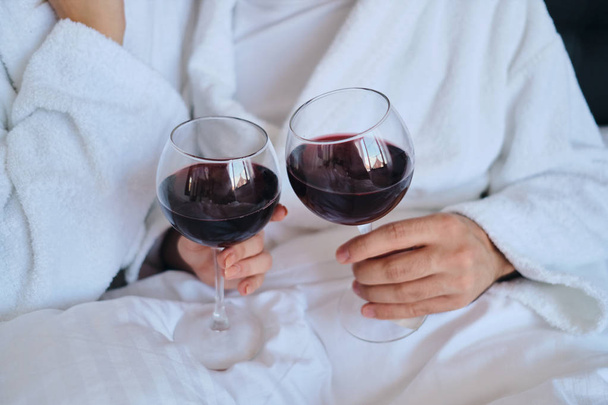 Close up man and woman in white bathrobes holding glasses of red wine in hands lying in bed in modern cozy hotel - Foto, Bild