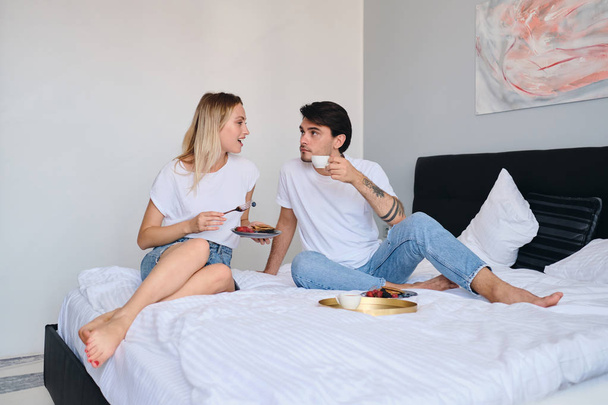 Young brunette man drinking coffee while pretty blond woman near eating pancakes with berries. Beautiful couple in white T-shirts having breakfast in bed in modern cozy hotel - Foto, Imagem