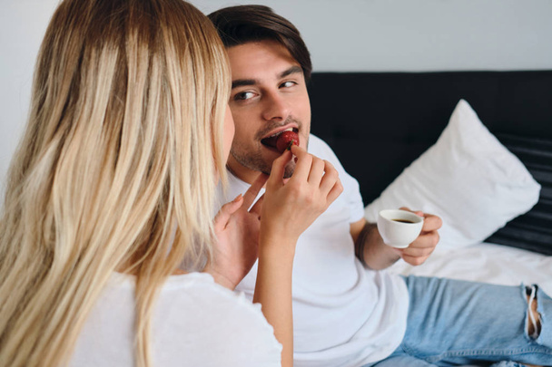 Young brunette man holding cup of coffee in hand dreamily eating strawberry and looking at blond woman feeding him. Couple in white T-shirts having breakfast in bed in modern hotel - Photo, Image