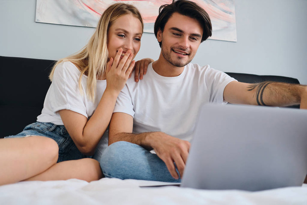 Beautiful blond woman and smiling brunette man happily watching movie on laptop together. Young beautiful couple in white T-shirts sitting on bed at cozy home - Fotografie, Obrázek