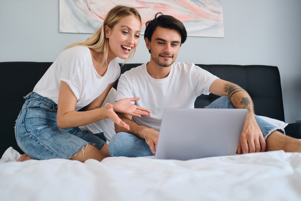 Femme blonde joyeuse et homme brune souriant heureux de travailler sur un ordinateur portable ensemble. Jeune beau couple en T-shirts blancs assis sur le lit à la maison confortable
 - Photo, image
