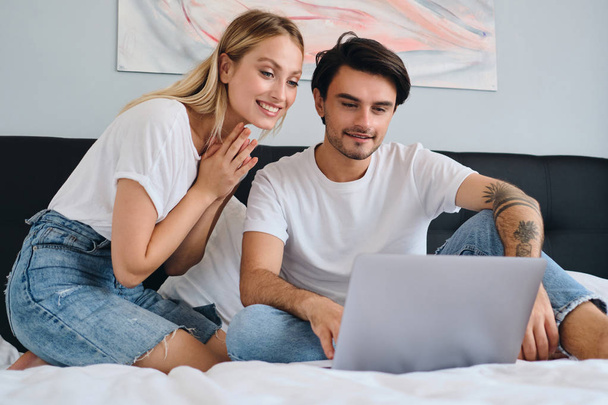 Όμορφη ξανθιά γυναίκα και ελκυστική μελαχρινή άντρας που εργάζονται μαζί στο laptop μαζί. Νεαρό όμορφο ζευγάρι σε λευκά μπλουζάκια που κάθονται στο κρεβάτι στο άνετο σπίτι - Φωτογραφία, εικόνα