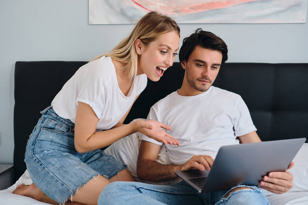 Αρκετά συναισθηματική ξανθιά γυναίκα που κοιτάζει χαρούμενα στο laptop, ενώ ελκυστική μελαχρινή άνθρωπος που εργάζονται σε αυτό. Νεαρό όμορφο ζευγάρι με λευκά μπλουζάκια που κάθονται μαζί στο κρεβάτι στο άνετο σπίτι - Φωτογραφία, εικόνα