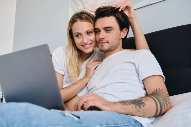 Χαρούμενη ξανθιά γυναίκα αγκαλιάζοντας ελκυστικό μελαχρινή άνθρωπος, ενώ ονειρικά εργάζεται σε φορητό υπολογιστή. Νεαρό όμορφο ζευγάρι με άσπρα μπλουζάκια που κάθονται στο κρεβάτι μαζί στο σπίτι - Φωτογραφία, εικόνα