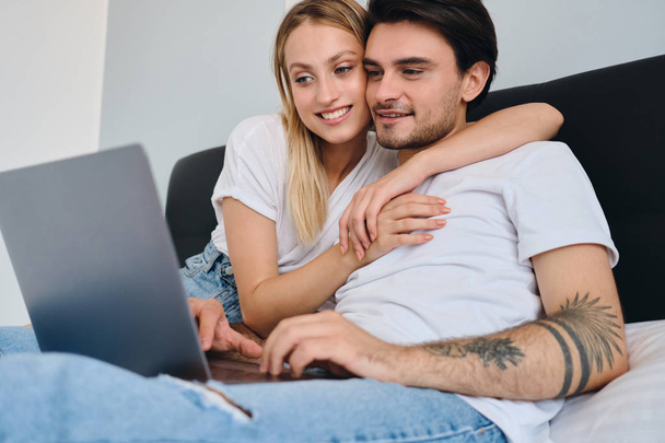 Όμορφη ξανθιά γυναίκα αγκαλιάζει ευτυχισμένοι ελκυστικό μελαχρινή άνθρωπος, ενώ εργάζεται για το laptop. Νεαρό όμορφο ζευγάρι με άσπρα μπλουζάκια που κάθονται στο κρεβάτι μαζί στο σπίτι - Φωτογραφία, εικόνα