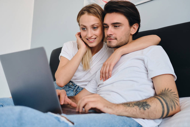 Atrakcyjny brunetka człowiek i piękny blond kobieta szczęśliwie przy pomocy laptop razem. Młoda piękna para w białych koszulkach siedzących na łóżku w domu - Zdjęcie, obraz