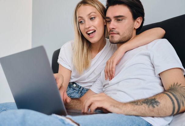 Young thoughtful brunette man and pretty emotional blond woman in white T-shirts using laptop together in bed at cozy home - Photo, Image