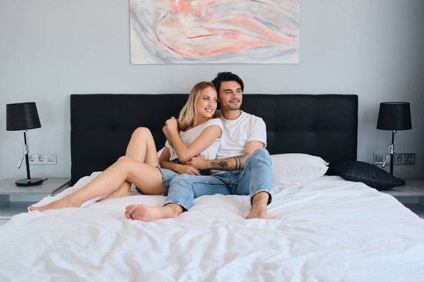 Beautiful blond woman and handsome brunette man hugging each other. Young attractive couple in white T-shirts lying on bed in modern cozy hotel happily looking aside together - Photo, Image