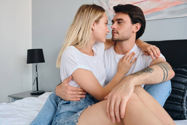 Handsome brunette man and pretty blond woman dreamily looking at each other and hugging. Young beautiful couple in white T-shirts sitting on bed together in modern cozy hotel - Foto, immagini