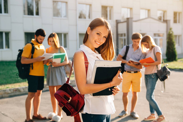 Красивая молодая девушка с красным бархатом рюкзак с книгами и улыбаясь, стоя против университета со своими друзьями на заднем плане
 - Фото, изображение