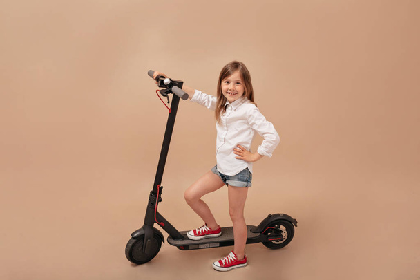 Indoor frame of little cute girl posing at camera over isolated beige background with electric scooter - 写真・画像