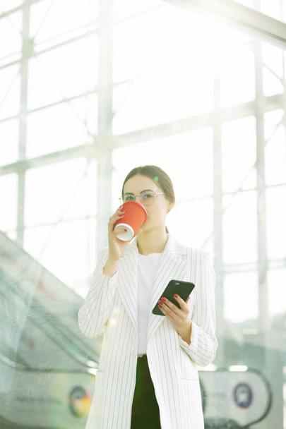 Cropped image of successful business woman wearing suit using modern smartphone and drinking coffee to go before going to work early in the morning - Фото, зображення