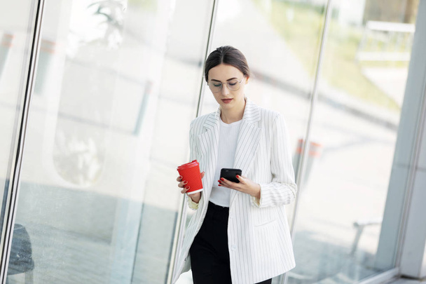 Cropped image of successful business woman wearing suit using modern smartphone and drinking coffee to go before going to work early in the morning - Фото, зображення