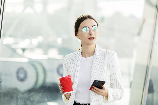 Cropped image of successful business woman wearing suit using modern smartphone and drinking coffee to go before going to work early in the morning - Fotoğraf, Görsel