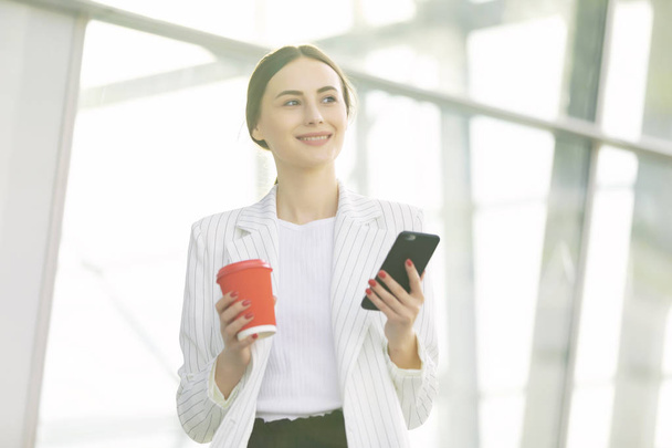 Cropped image of successful business woman wearing suit using modern smartphone and drinking coffee to go before going to work early in the morning - Valokuva, kuva