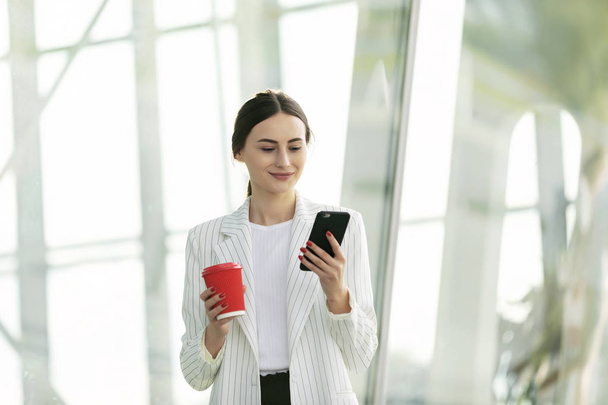 Cropped image of successful business woman wearing suit using modern smartphone and drinking coffee to go before going to work early in the morning - Foto, Bild