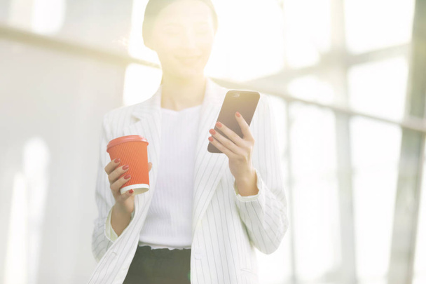 Cropped image of successful business woman wearing suit using modern smartphone and drinking coffee to go before going to work early in the morning - Zdjęcie, obraz