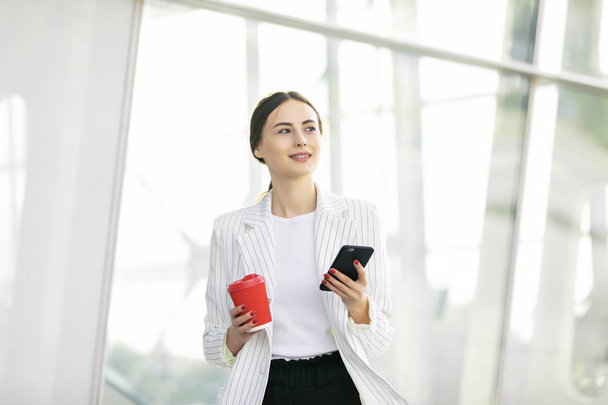 Cropped image of successful business woman wearing suit using modern smartphone and drinking coffee to go before going to work early in the morning - Foto, afbeelding