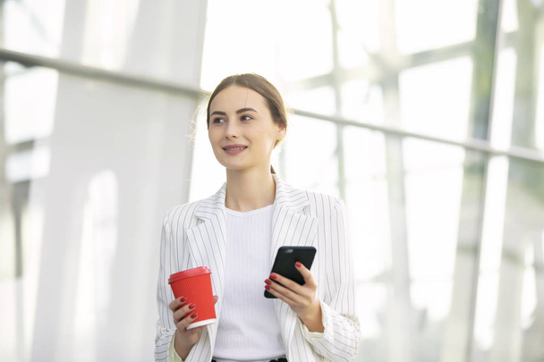 Cropped image of successful business woman wearing suit using modern smartphone and drinking coffee to go before going to work early in the morning - Zdjęcie, obraz