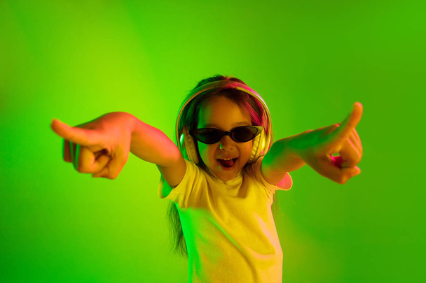 Portrait of little girl in headphones on green background in neon light - Photo, image