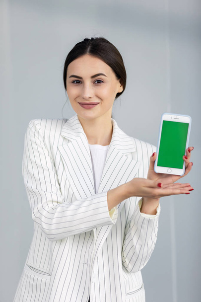 Smiling young business woman in white sweater showing smartphone with green screen, appSmiling young business woman in white sweater showing smartphone with green screen, app - Foto, immagini