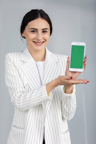 Smiling young business woman in white sweater showing smartphone with green screen, app - Fotografie, Obrázek