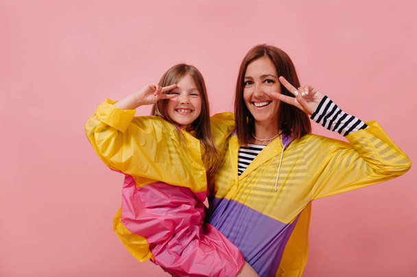 Studio shot of woman with little charming girl wearing raincoats showing peace signs over isolated pink background - Фото, изображение