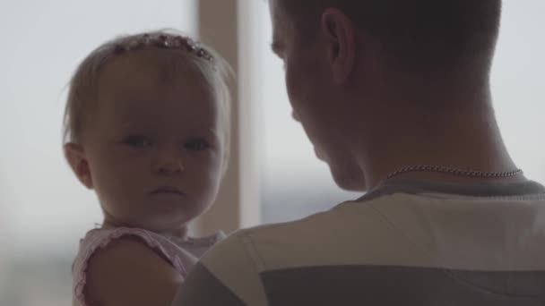 Handsome young man holding little baby girl in his arms in front of window close-up. The joys of fatherhood. Loving family at home. Beautiful child with father. Slow motion - Materiał filmowy, wideo