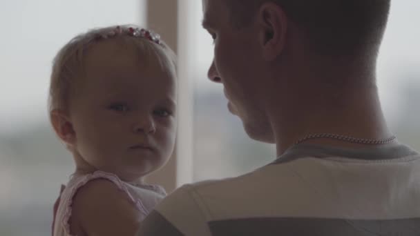 Handsome man holding little baby girl in his arms in front of window close-up. The joys of fatherhood. Loving family at home. Beautiful child with father. Slow motion - Filmati, video