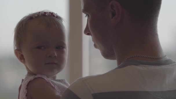Back view of the man holding little baby girl in his arms in front of window close-up. The joys of fatherhood. Loving family at home. Beautiful child with father. Slow motion - Кадри, відео