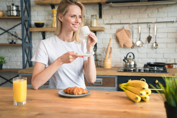 mooie jonge lachende vrouw zit in de keuken thuis, ontbijten, koffie drinken met croissants en zijdelings kijken. Ochtend huis, ontbijt. dragen van wit overhemd - Foto, afbeelding
