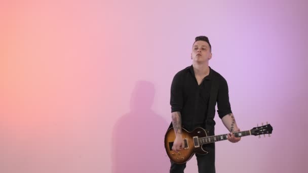 Young man in black shirt playing electric guitar with different colors at background. Rock musician plays solo guitar and looking at camera. Medium shot with copy space - Séquence, vidéo