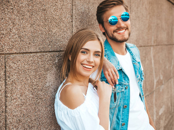 Smiling beautiful girl and her handsome boyfriend. Woman in casual summer dress.Man in jeans clothes.Happy cheerful family.Female having fun on the street near wall.Hugging couple in sunglasses - Foto, Imagem
