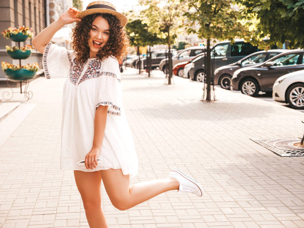 Portrait of beautiful smiling model with afro curls hairstyle dressed in summer hipster white dress.Sexy carefree girl posing in the street background.Trendy funny and positive woman in hat - 写真・画像
