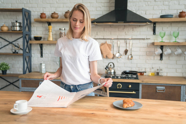 joven hermosa mujer está leyendo su prensa de la mañana mientras desayuna en la cocina. café con croissants. con camisa blanca y jeans
 - Foto, Imagen