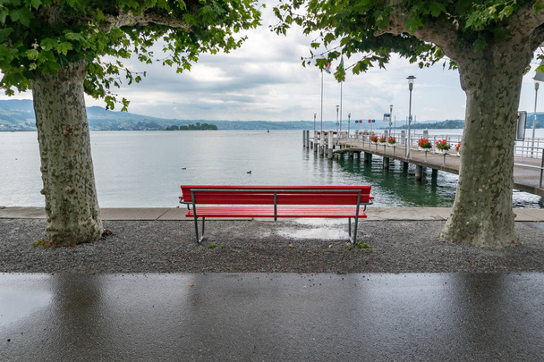 red park bench on a lake shore with a view of the harbor pier an - Photo, Image