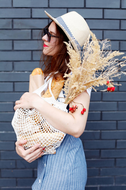 beautiful stylish girl in a linen sundress, a straw hat, sunglasses and a white T-shirt in sneakers with a cotton shopping bag, baguette and dry flowers of a gray brick wall. Eco friendly - Foto, Bild