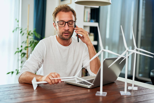 Handsome Caucasian ecology engineer sitting at home, talking on the phone about project and holding windmill model . On desk are windmill models. Sustainable development concept. - Foto, Imagen