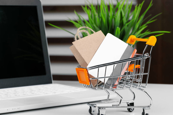 Online shopping concept. Shopping cart, laptop on the desk - Фото, изображение