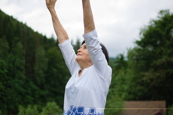 Donna anziana che fa un esercizio di stretching per la parte superiore delle braccia al di fuori sul paesaggio della foresta e delle montagne
 - Foto, immagini