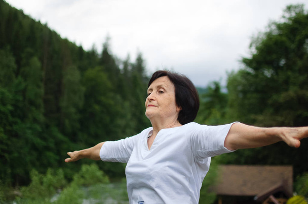 Starsza kobieta robi ćwiczenia rozciągające dla górnych ramion poza krajobraz lasu i gór - Zdjęcie, obraz