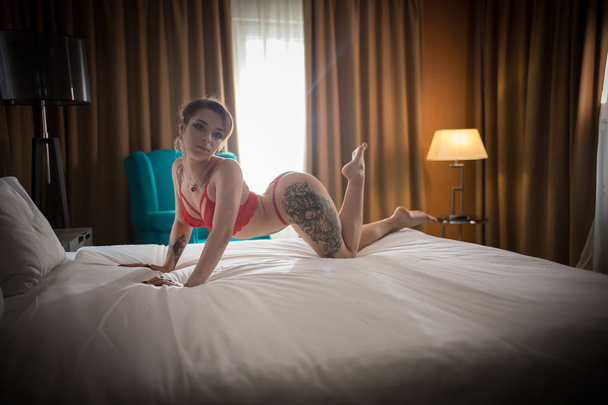 Young woman with tattoos in red underwear gracefully standing on the knees on the bed - Фото, изображение