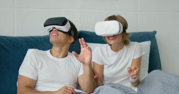 Young mixed couple lying in bed, watching 360 video in virtual reality headsets - Záběry, video