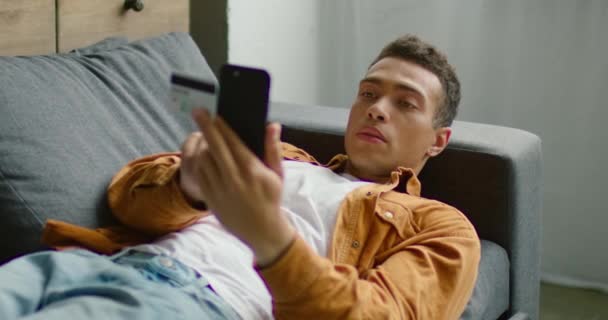 Hispanic handsome man is lying on sofa, holding smartphone and credit card - Felvétel, videó