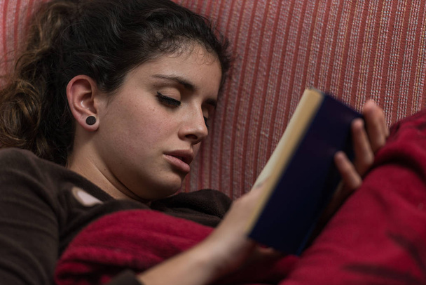 茶色の髪をした若い女性は、本を読んでいる間にソファで眠りに落ちる. - 写真・画像