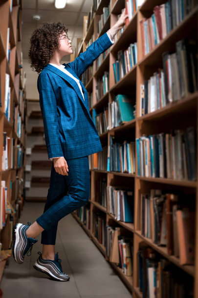 Young beautiful curly girl in glasses and a blue suit is standing in the library - Foto, imagen
