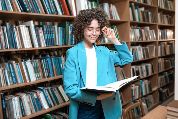 Young beautiful curly girl in glasses and a blue suit is standing in the library - Photo, Image