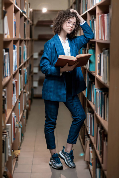 Młoda piękna dziewczyna kręcone w okularach i niebieski garnitur stoi w bibliotece - Zdjęcie, obraz