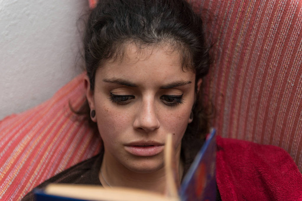 Νεαρή γυναίκα με καστανά μαλλιά διαβάζει ήσυχα ένα βιβλίο που βρίσκεται στον καναπέ τυλιγμένο σε μια κόκκινη κουβέρτα. - Φωτογραφία, εικόνα