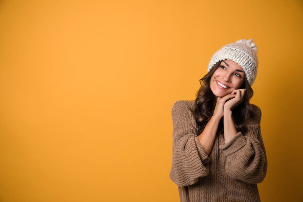 Young woman in warm sweater and hat on yellow background, space for text. Christmas season - Фото, изображение